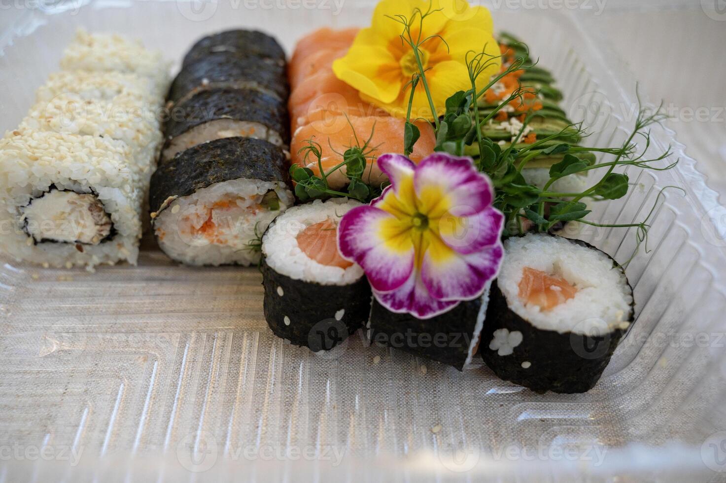 Food delivery concept. Transparent plastic lunch box with sushi set. Mixed sushi rolls with salmon, avocado, smoked eel, sesame in delivery box. sushi decorated with flowers. on natural tablecloth background on table. photo