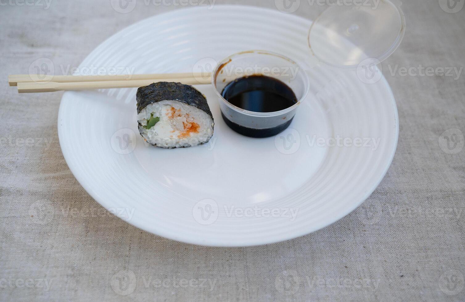 sushi roll with chopsticks and wasabi sauce in a plastic container on a white plate. elements of japanese cuisine, takeaway food. linen tablecloth on the table. photo