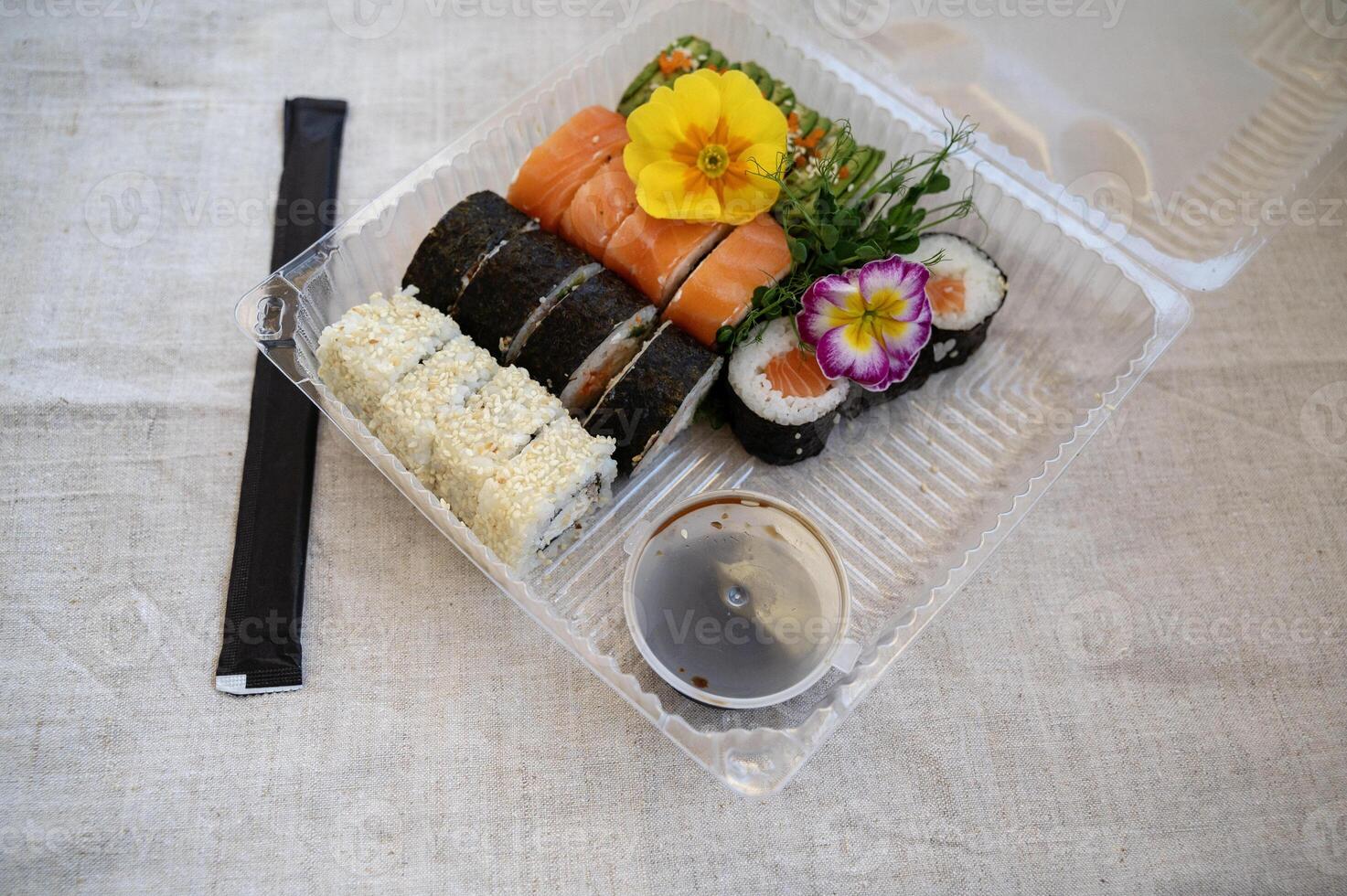 Food delivery concept. Transparent plastic lunch box with sushi set. Mixed sushi rolls with salmon, avocado, smoked eel, sesame in delivery box. Top view of sushi assortment on gray natural tablecloth on table. Takeaway food photo