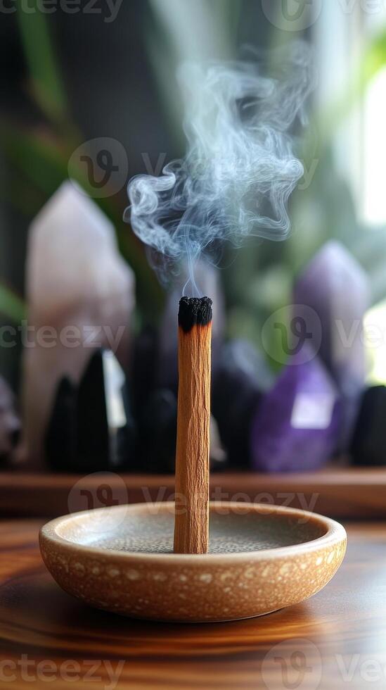 An incense palo santo stick emitting smoke, in round ceramic holder on a wooden surface. Healing crystals in background photo