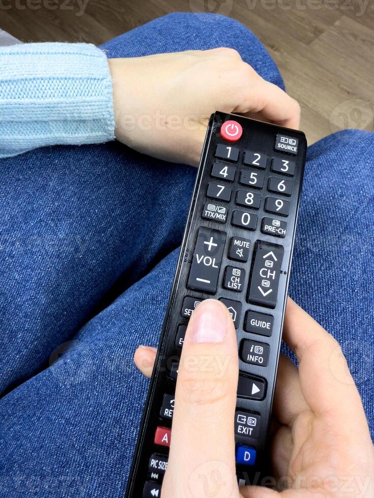 The TV remote control in hands of girl lies on her legs while watching an interesting television program. Control your TV at home using remote controller, vertical photo of TV controller