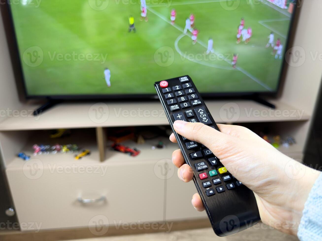 Girl holds remote control in her hand and make sound higher while watching TV with football match. Girl watches football on TV. Watching an online broadcast of football match while lying on sofa photo