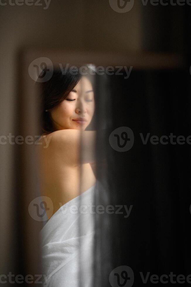 Wrapped in a towel, an Asian womans delicate reflection appears softly in the mirror photo