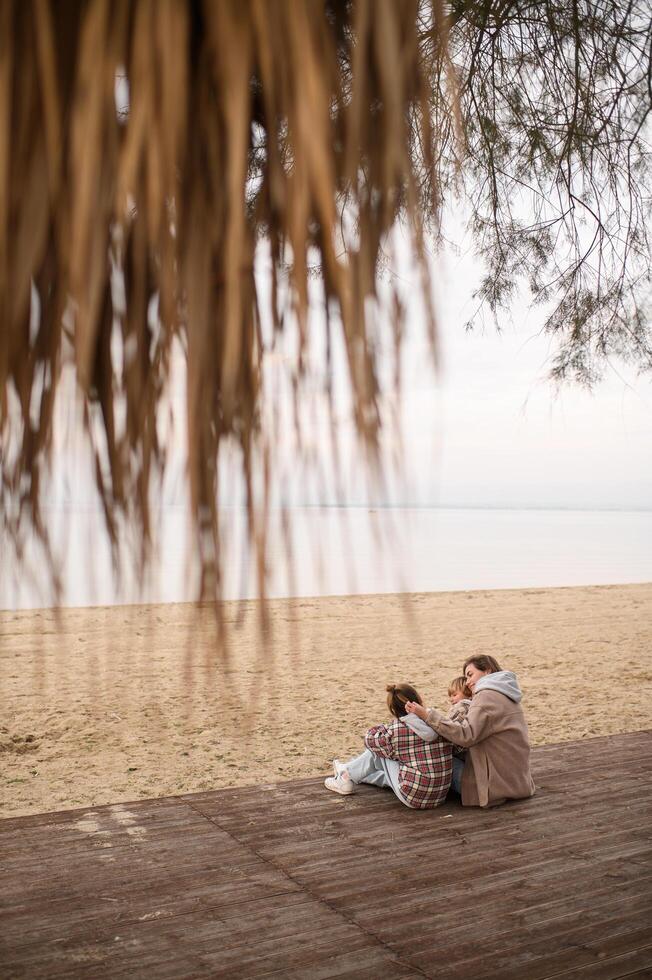 The family sits together on a wooden platform near the shore, sharing warmth and love on a peaceful fall afternoon photo
