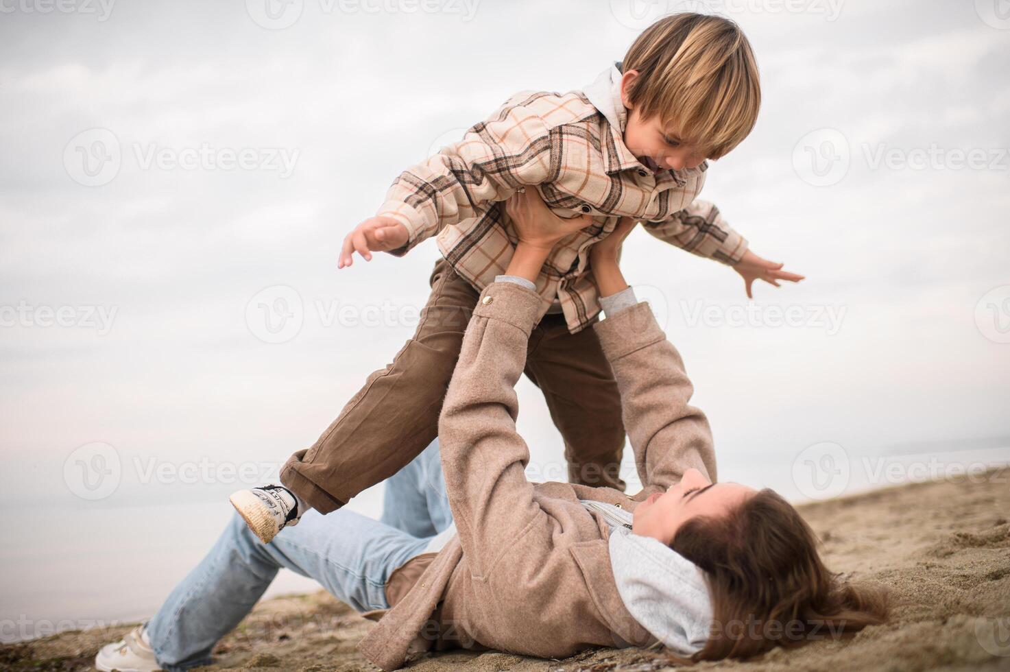 A mother plays with her baby boy on the beach, holding him above her as he spreads his arms like an airplane photo
