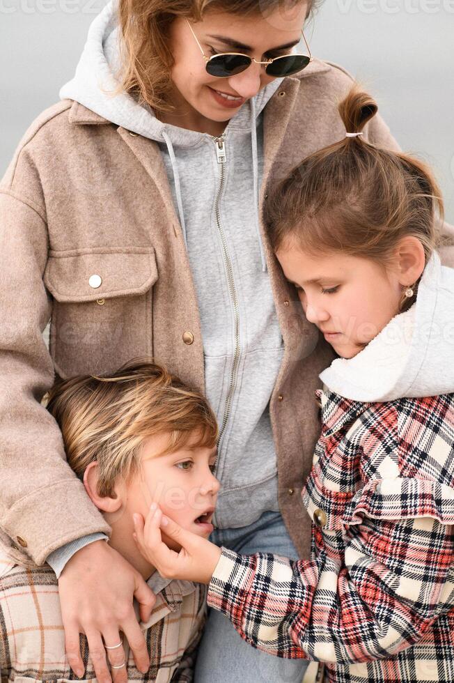 A close-up portrait of a mother in glasses hugging her children as they play together in their plaid jackets on a crisp autumn day. photo