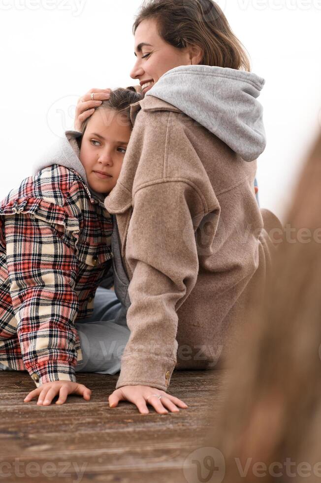 The mother gently hugs her daughter as they sit together on a wooden deck near the shore, enjoying the fresh sea breeze. photo