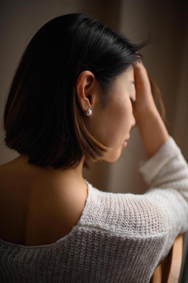A young Asian woman gazes softly, wearing a cozy knit sweater and an elegant pearl earring. photo