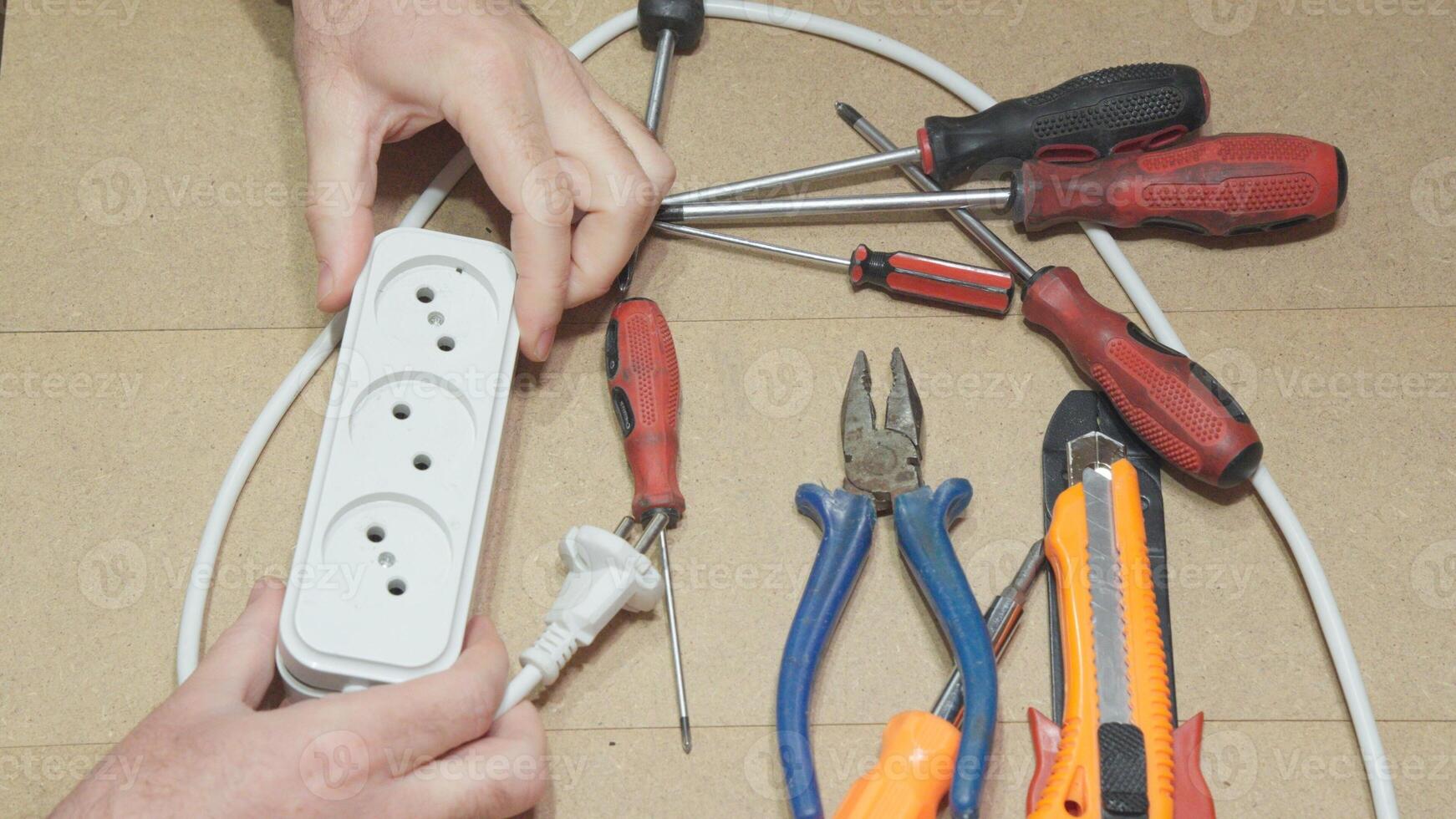 A hand holds an extension cord next to tools screwdrivers, pliers, and a knife. photo