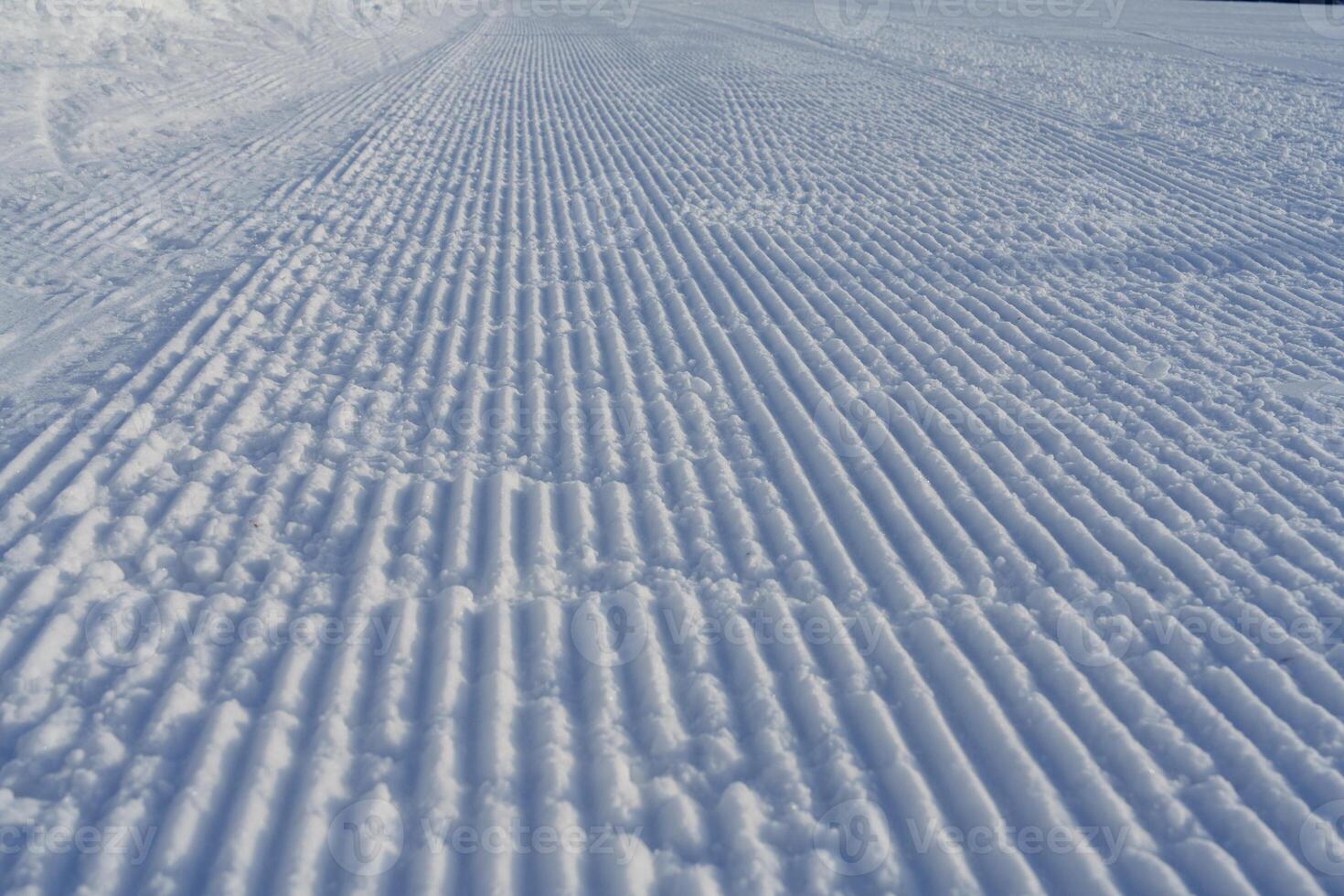 A groomed snow track at a ski resort features fresh white snow and clear track marks from skiers and snowboarders, capturing the thrill of winter sports in a picturesque landscape. photo