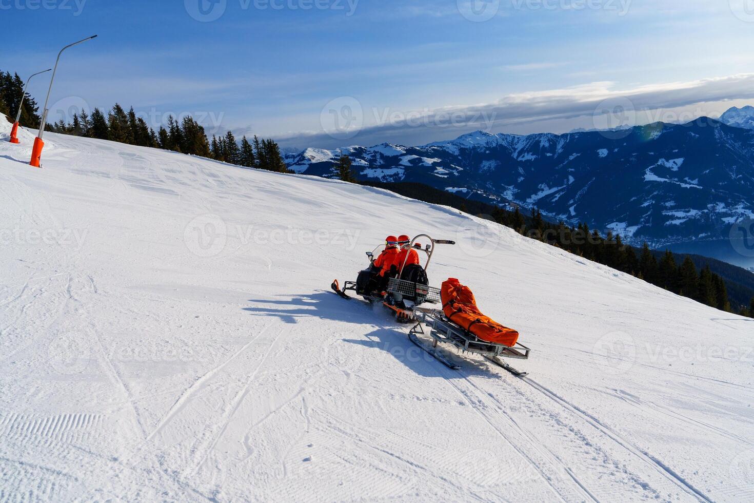 A snowmobile rescuer is driving a rescue sled with an injured skier on it. The background is a beautiful mountain landscape with snow-covered trees and mountains. photo