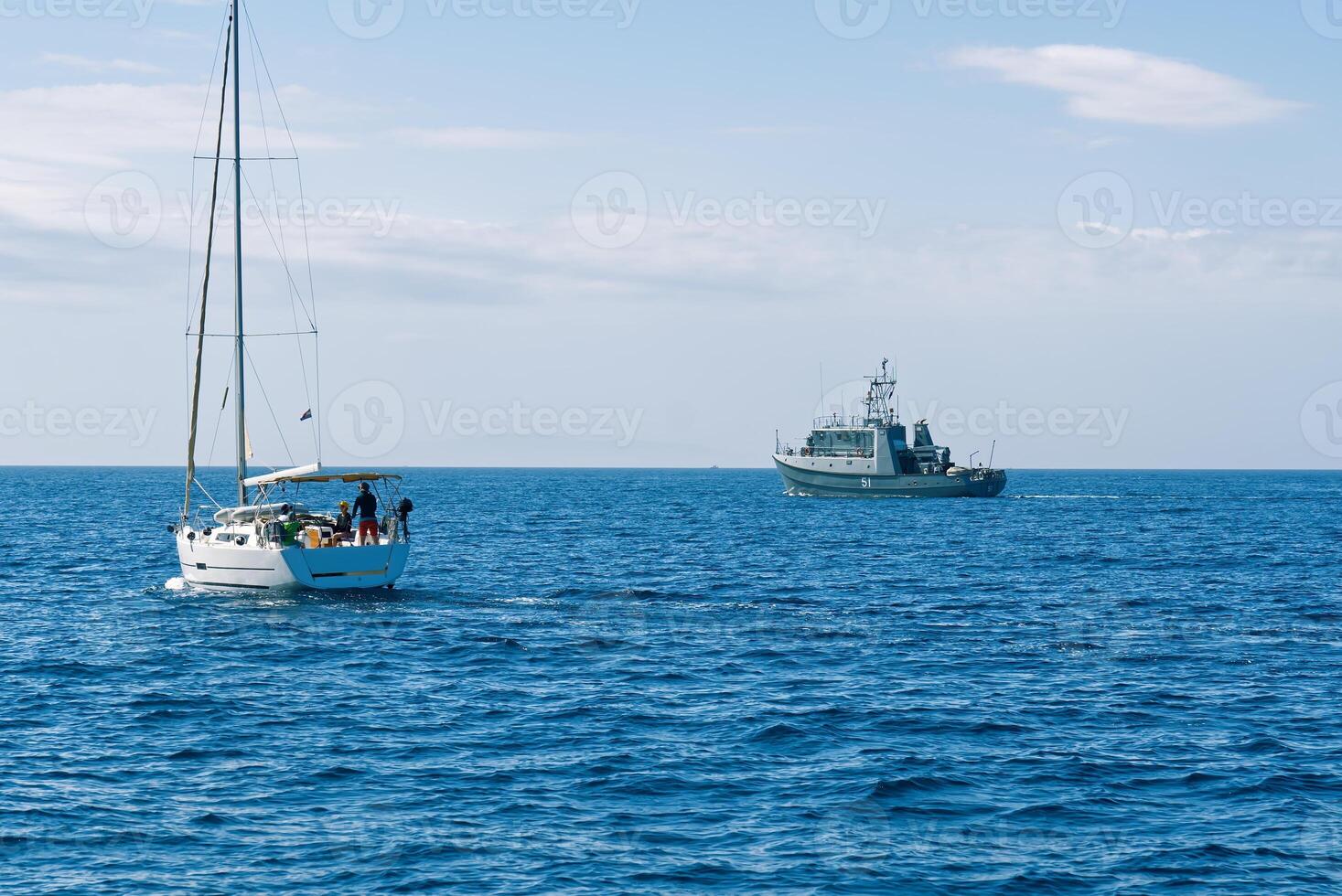 A small sailboat with three individuals is being inspected by an armed forces vessel in the open sea, showcasing maritime security operations and the importance of safeguarding waters. photo