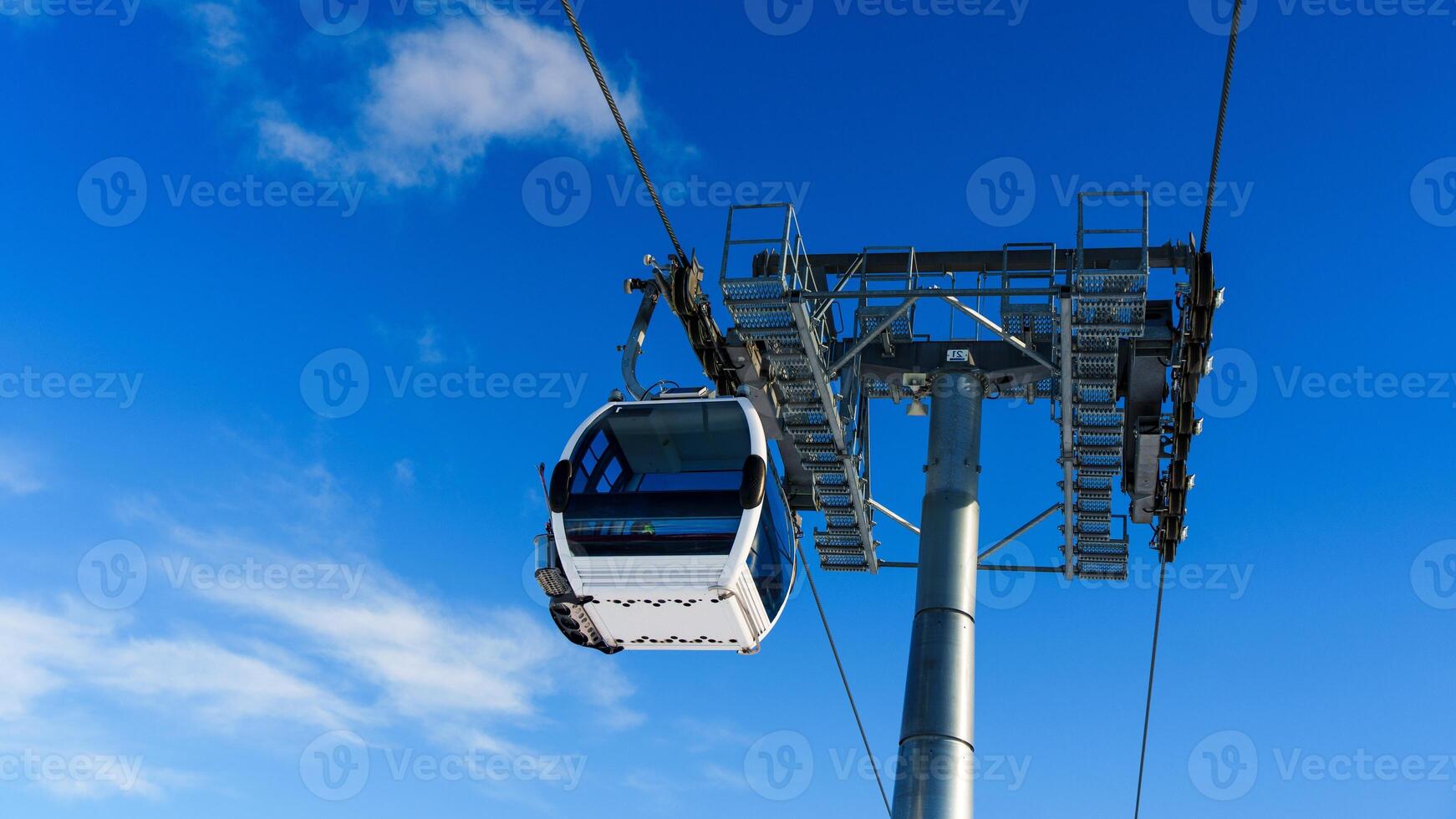 A modern white cable car is suspended from a cable in a clear blue sky. The sleek design and two supporting towers make it ideal for travel and tourism-related applications. photo
