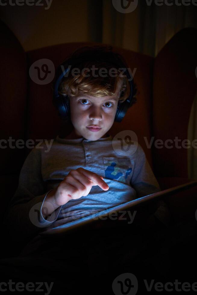 Vertical photo of a child sitting with helmets on a sofa touching the screen of an ipad and looking frontally at night
