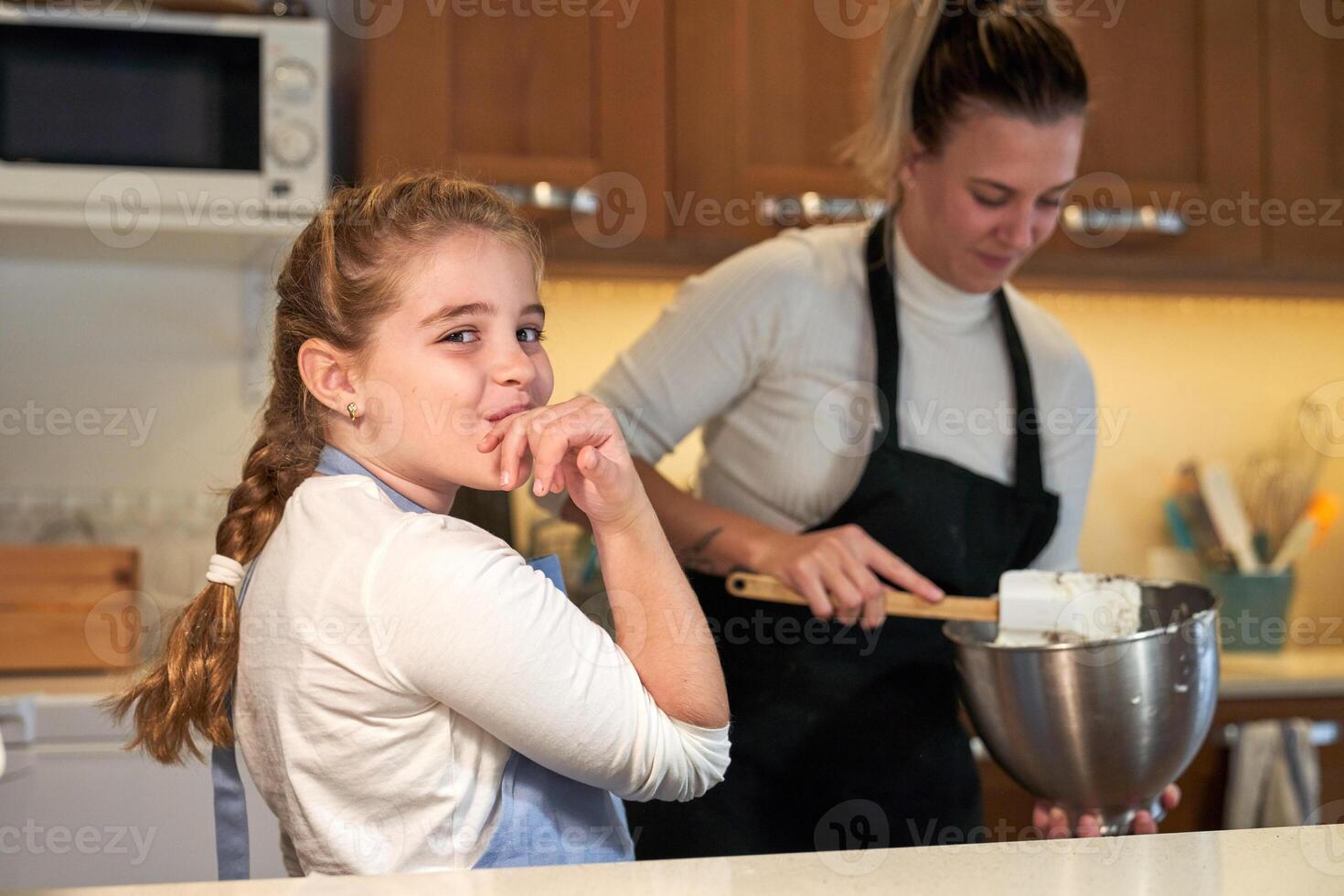 Girl in apron licking her hand in the kitchen with a girl in the background mixing ingredients in a metal container photo