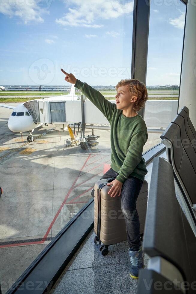 Happy kid pointing away sitting near window in airport terminal photo
