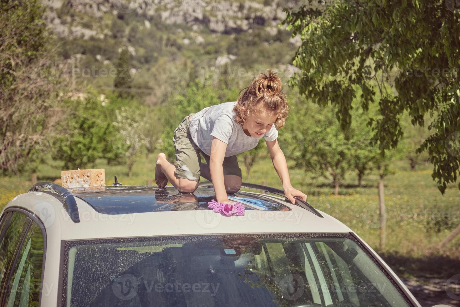 Focused kid with wash cloth and sponge on car roof in nature photo