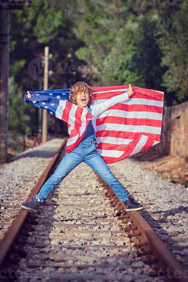 Joyful boy standing with USA flag on rails of railway photo