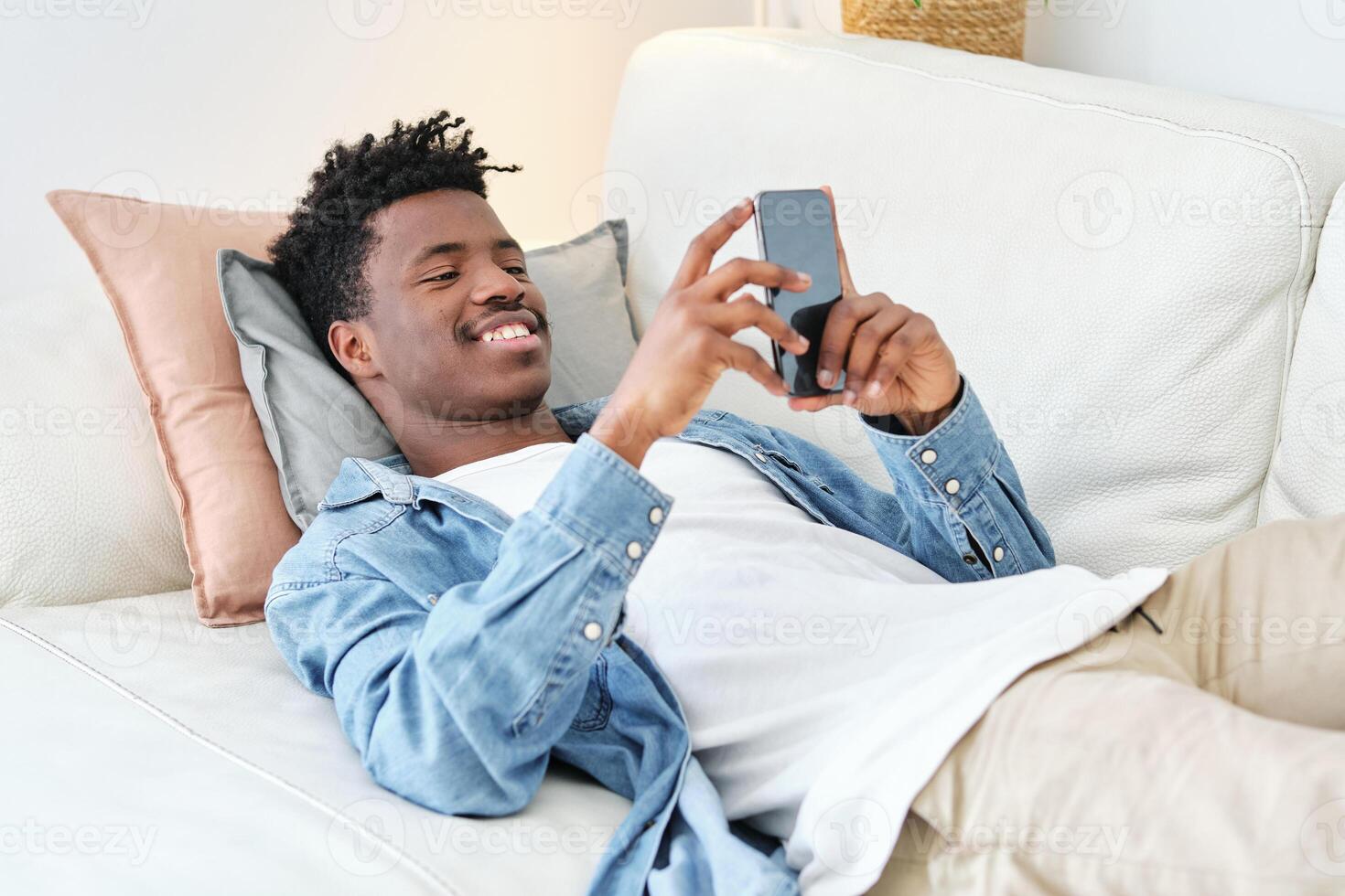 Happy black man lying on sofa and using smartphone photo