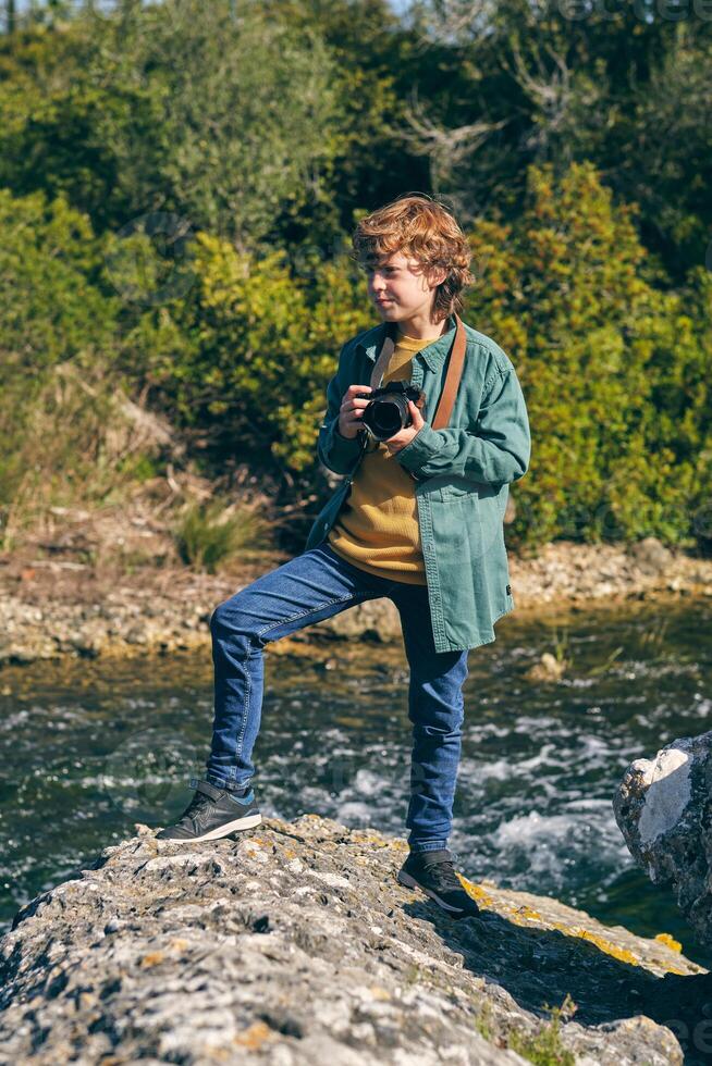 Content boy with photo camera standing on stone near brook