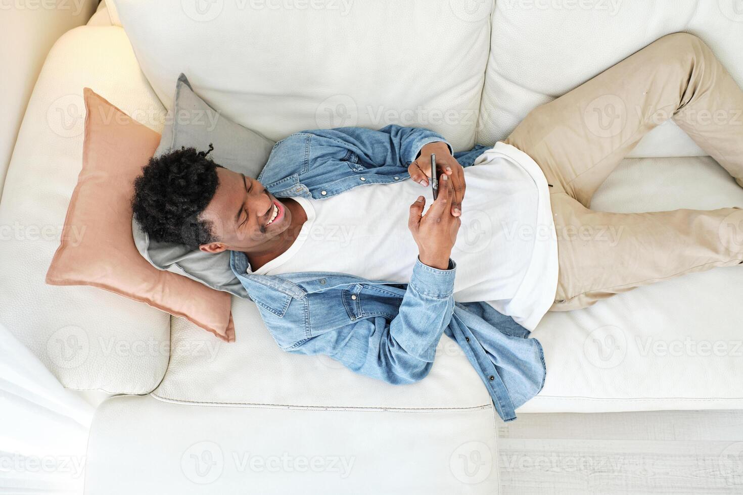 Happy black man resting on sofa and using smartphone photo
