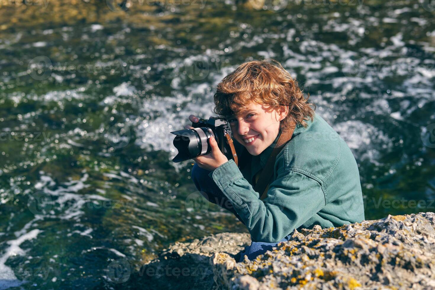 Smiling boy taking photo on rocky shore near stream of water