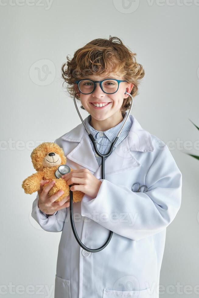 Cheerful boy in medical uniform examining toy bear with stethosc photo