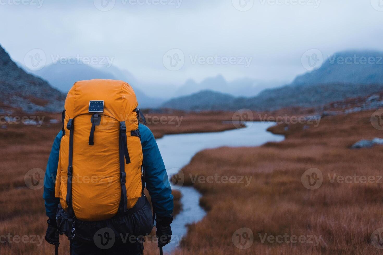 Mountains loom in the distance as a hiker with a yellow backpack gazes over a river under a moody sky in autumn photo