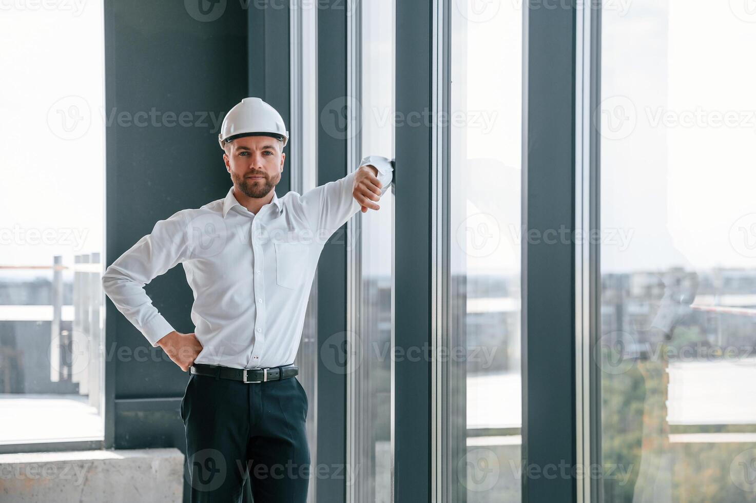 Standing near ,panoramic windows. Man in white shirt is working indoors at construction site. Managing the project photo
