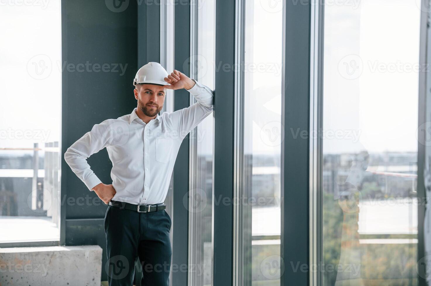 Standing, near panoramic windows. Man in white shirt is working indoors at construction site. Managing the project photo