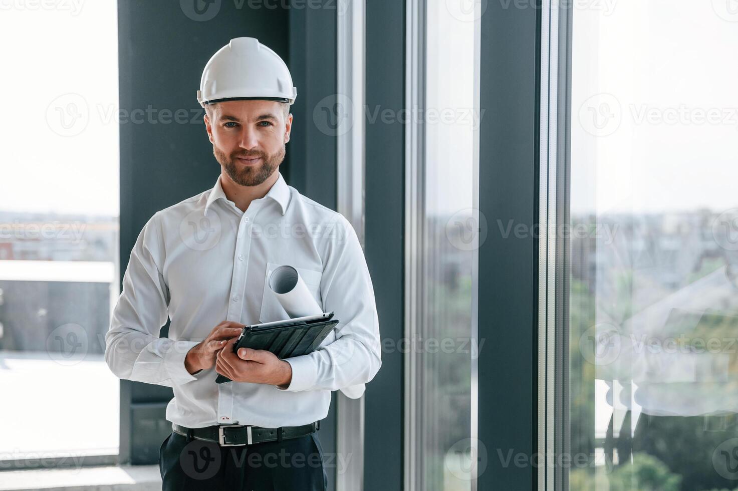 Standing near panoramic windows. Man in white shirt is working indoors at construction site. Managing the project photo
