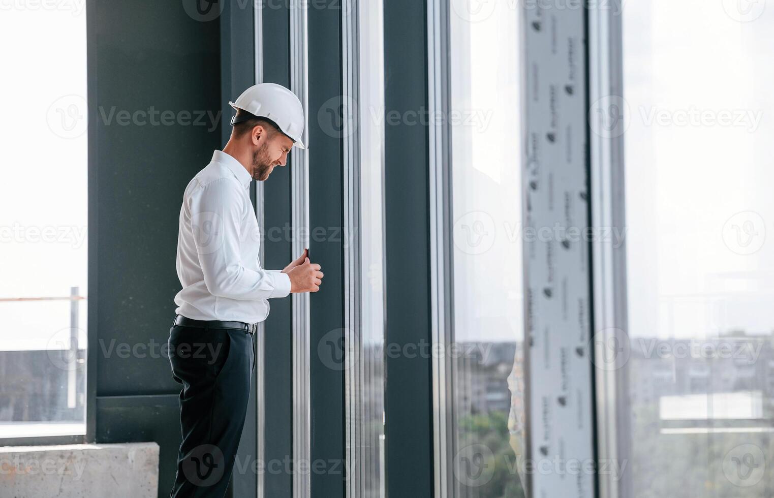 Standing ,near panoramic windows. Man in white shirt is working indoors at construction site. Managing the project photo