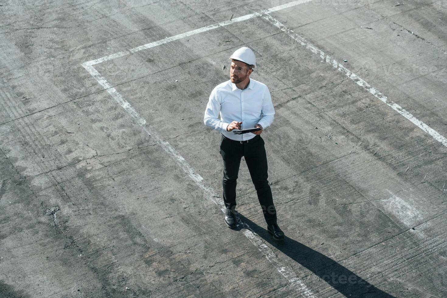 Top view of man in formal clothes and in white hard hat that is walking with tablet in hands photo