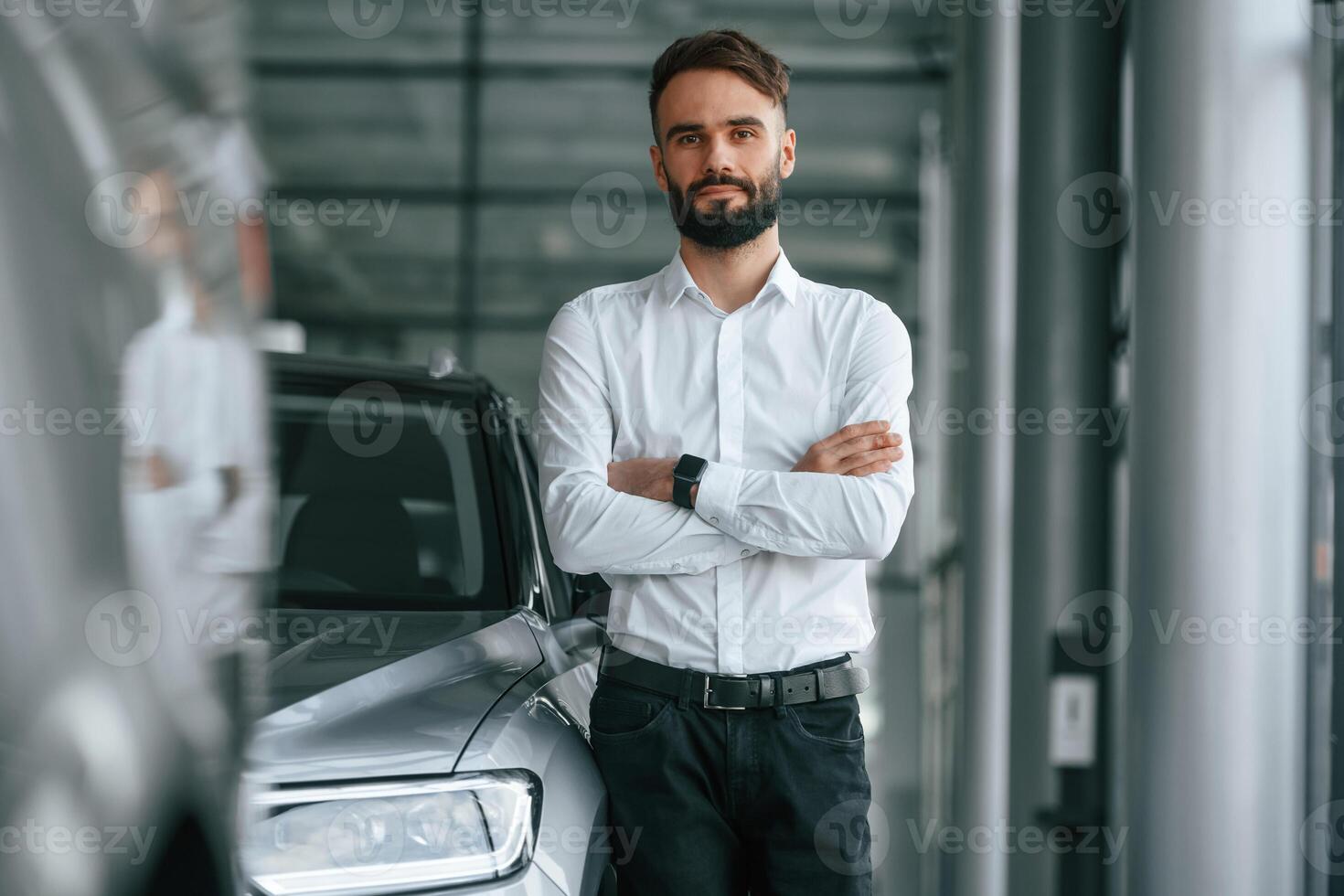Standing ,with arms crossed. Young man in white clothes is in the car dealership photo