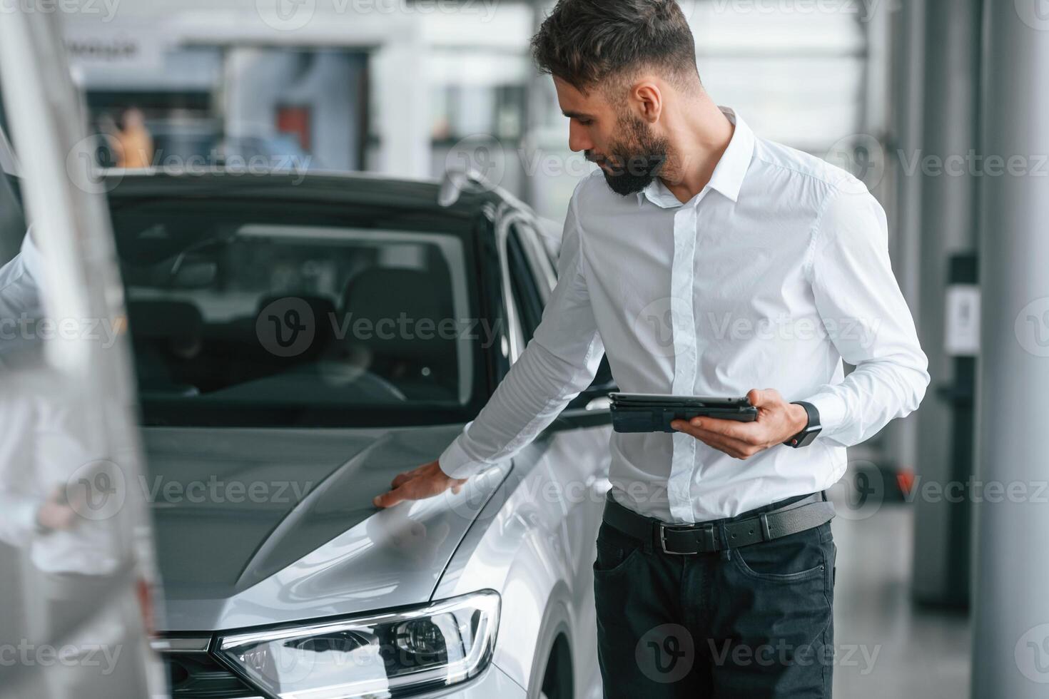 Touching the surface of automobile. Young, man in white clothes is in the car dealership photo