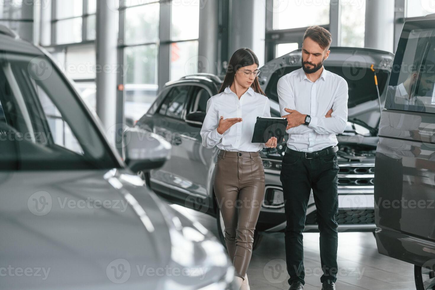 Reading ,information by using, tablet. Man with woman in white clothes are in the car dealership together photo