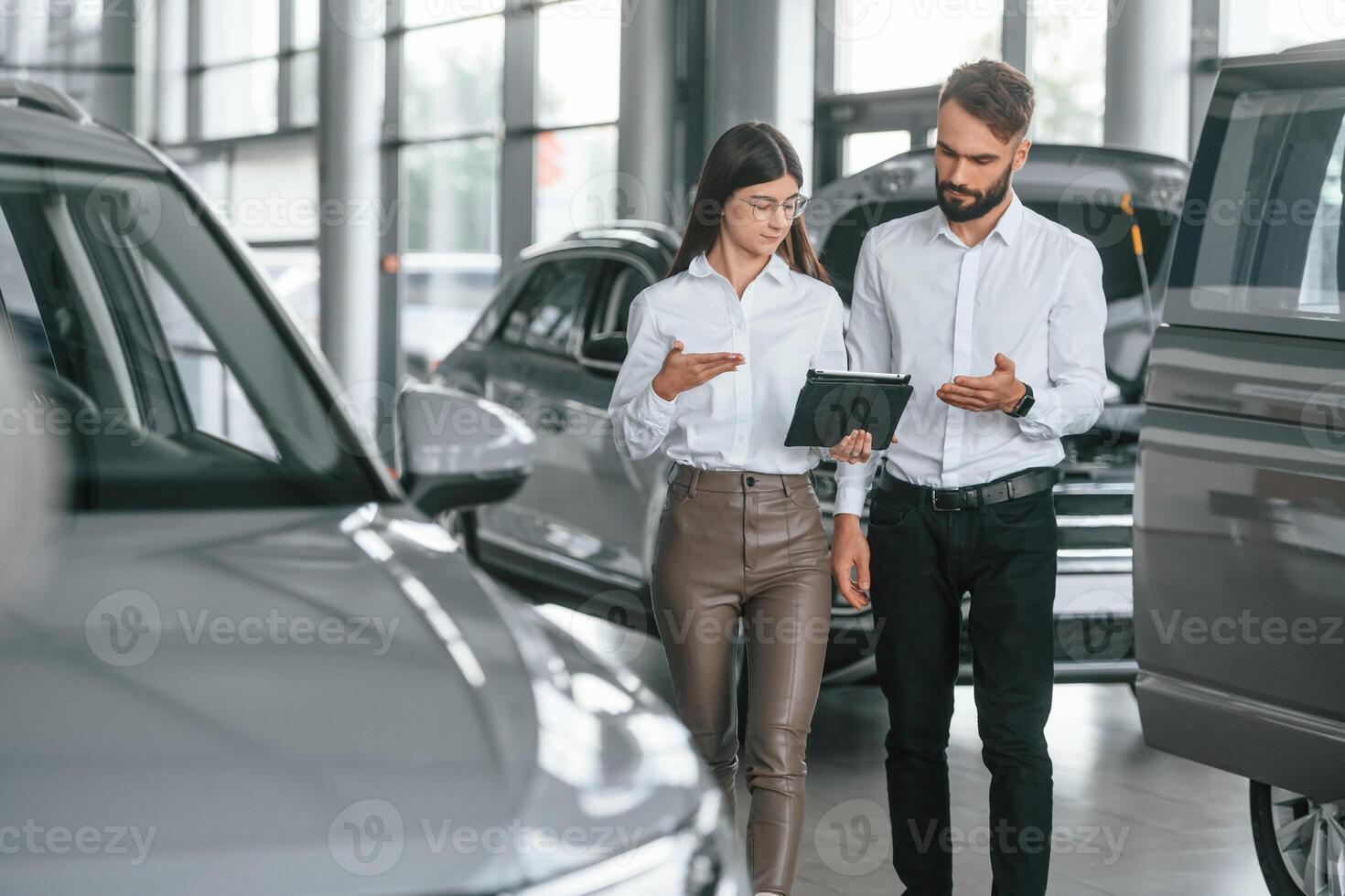 Reading information by using tablet. Man with woman in white clothes are in the car dealership together photo