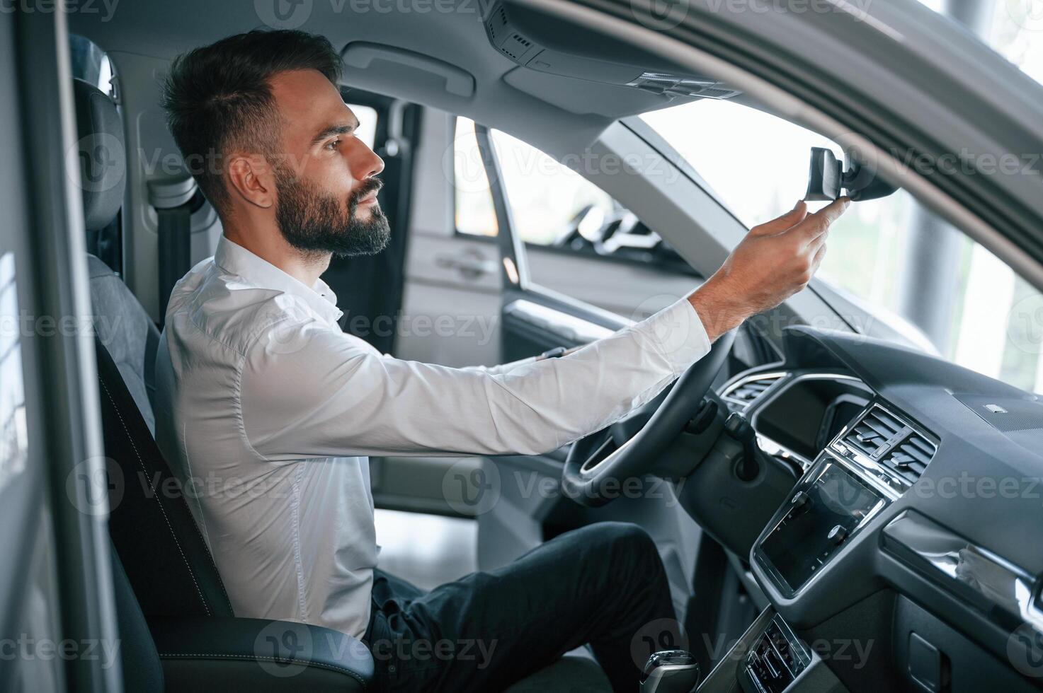 Side view. Sitting i,nside of a automobile. Young man in white clothes is in the car dealership photo