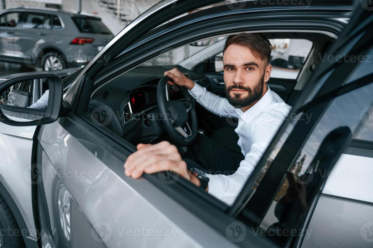 Sitting, looking ,through the window of the automobile. Young man in white clothes is in the car dealership photo