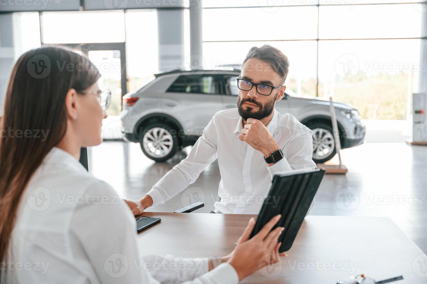 Showing information in the tablet. Sitting, by ,the table. Man with woman in white clothes are in the car dealership photo