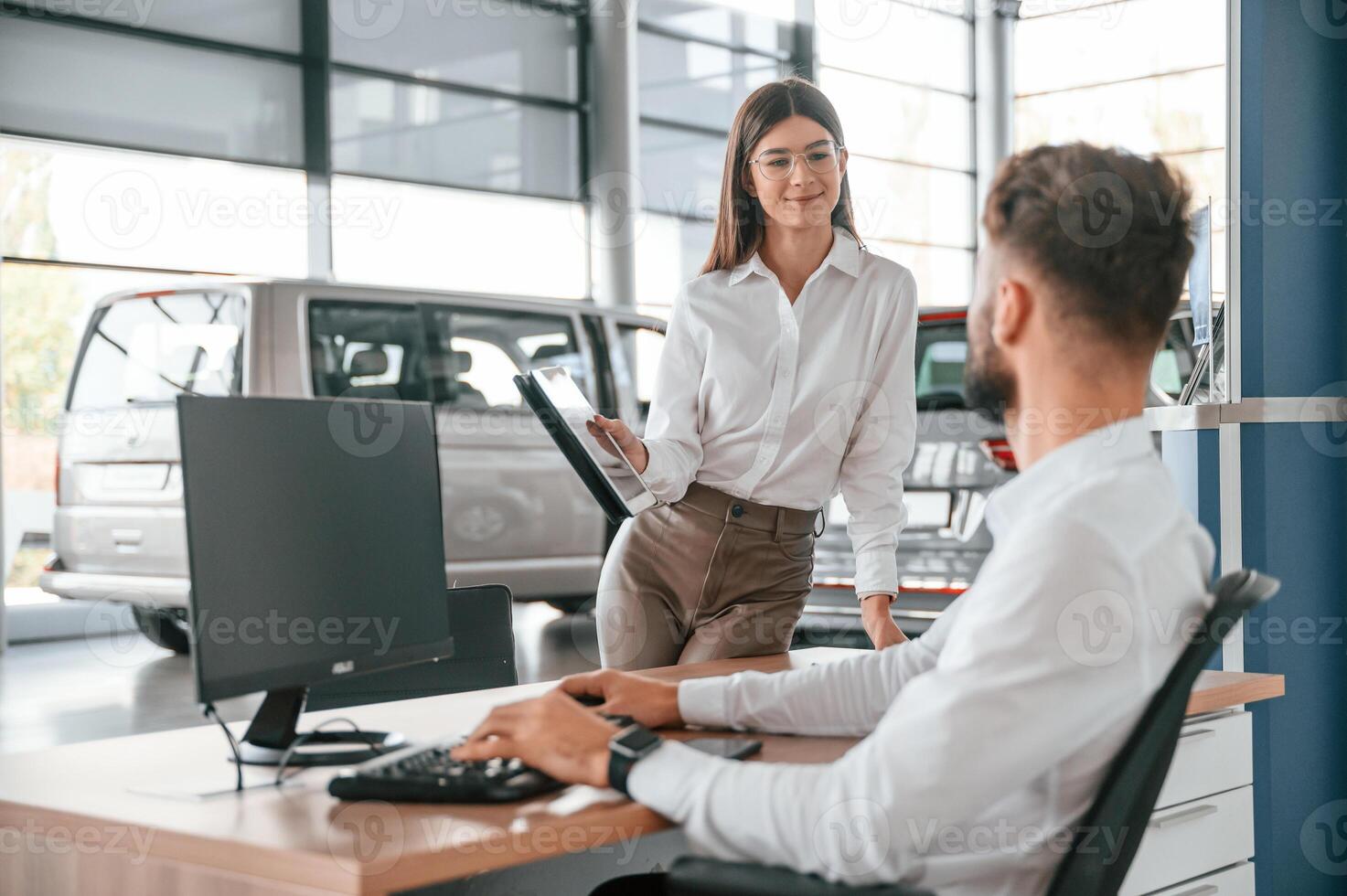 Sitting by a computer.. Man with woman in white clothes are in the car dealership together photo