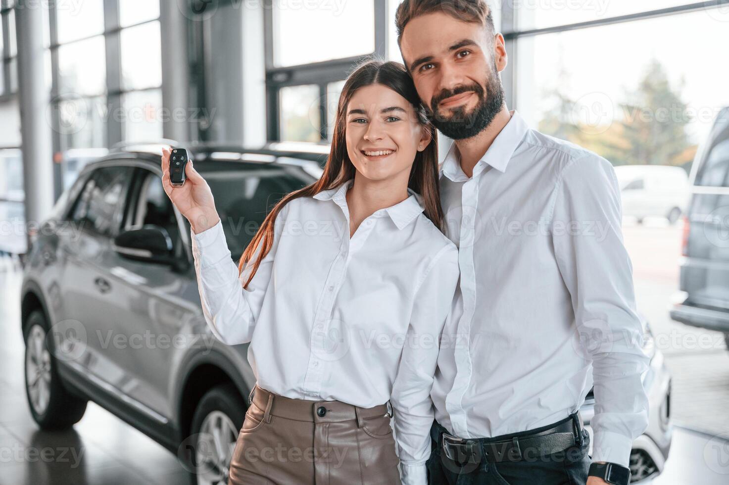 Standing with keys in hands. Happy ,owners.. Man with woman in white clothes are in the car dealership together photo