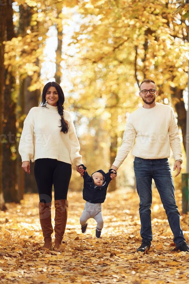 Holding toddler by the hands. Two parents are ,having a walk with their little son in the autumn park photo