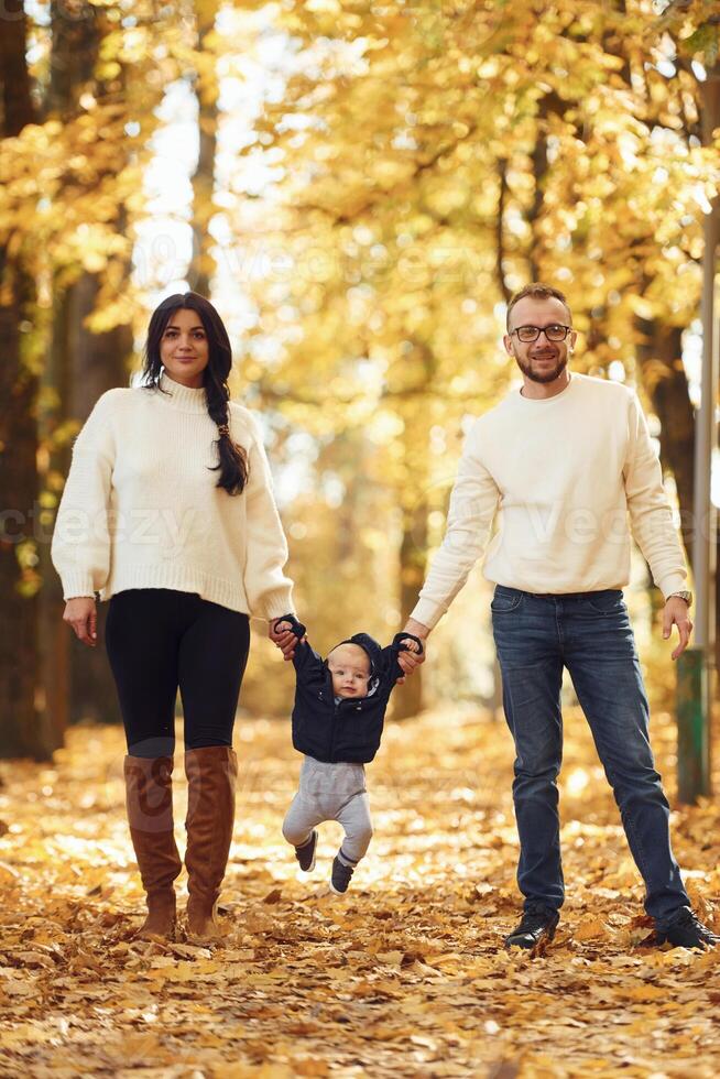 Holding toddler by, the hands. Two parents are having a walk with their little son in the autumn park photo