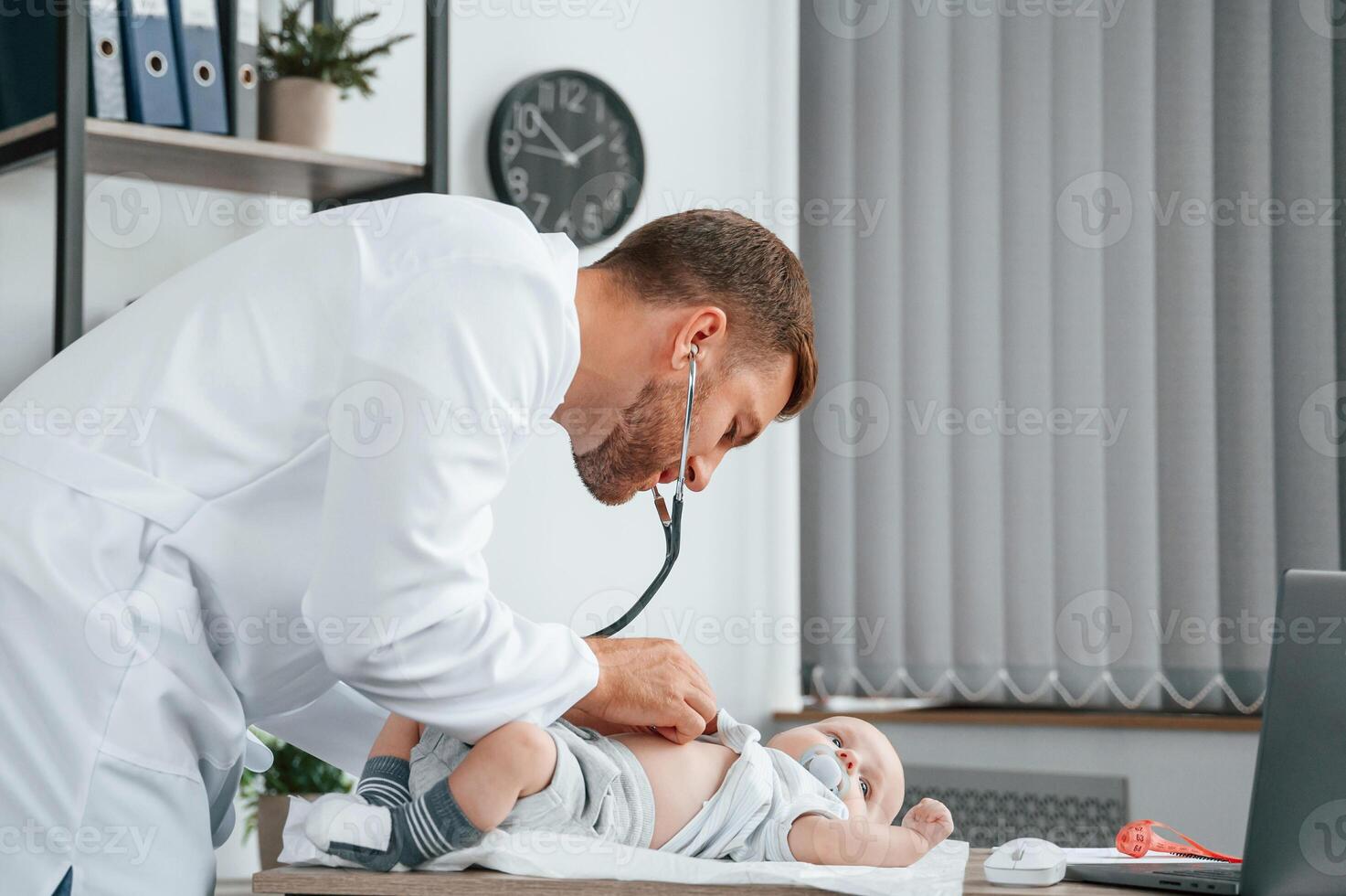 Using ,stethoscope. Doctor in white coat is taking care of little toddler indoors photo