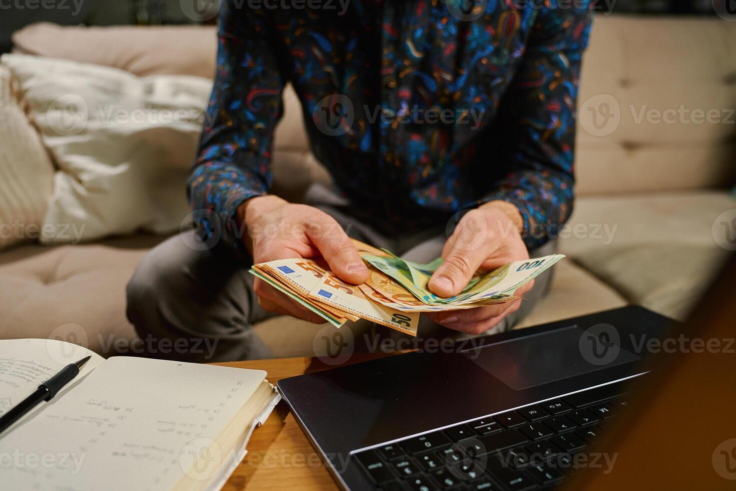 Man holding euro banknotes, sitting on sofa in front of table with laptop and notebook. Person counting remaining money after paying bills and utility expenses. Personal finance and home budget photo