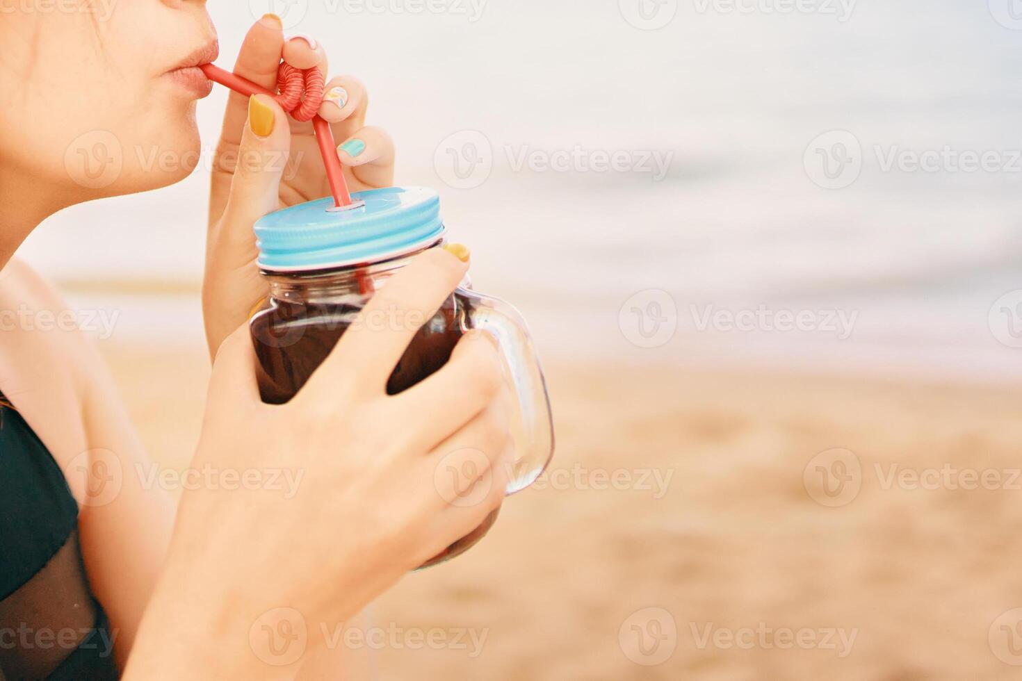 Close-up of a woman's lips drinking alcoholic cocktail with a straw. Sea beach in the background. Lady on vacation in the tropics. Photo card with copy space.