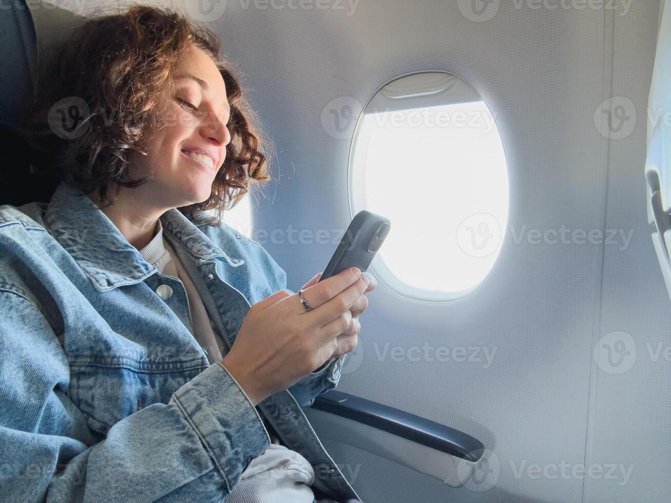 Passenger woman is flying in plane. Girl using phone sitting by airplane window. Traveling female with smartphone inside plane enjoying flight. Woman using wifi connection on board. Traveling girl photo