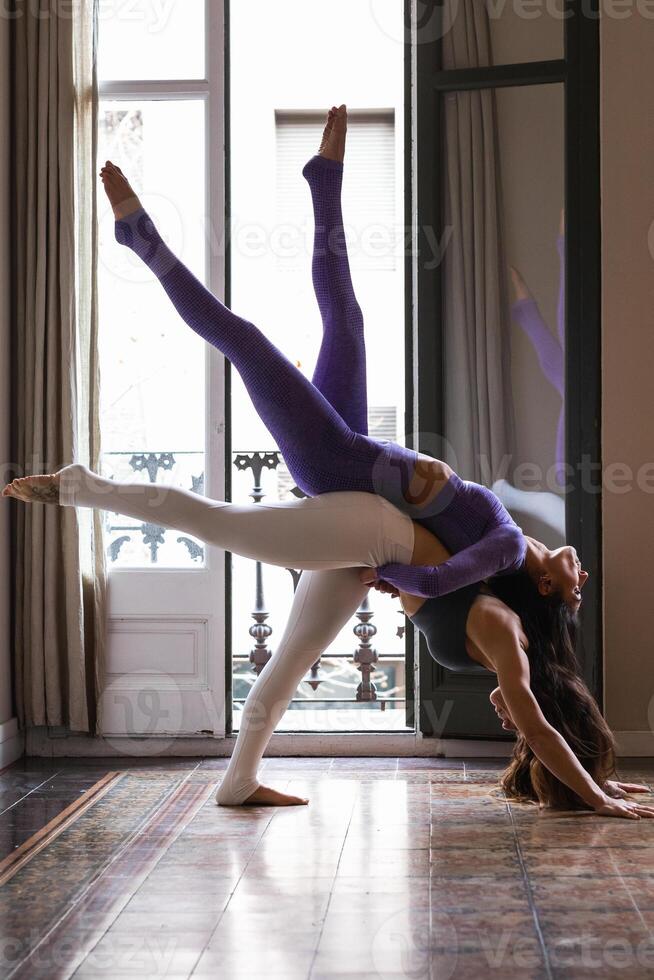 Flexible woman supporting female friend on back while doing Eka Pada Adho Mukha Svanasana yoga pose photo