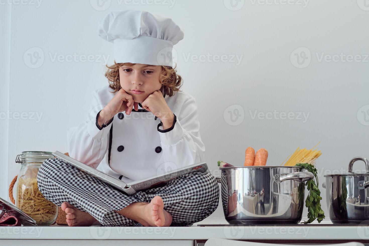 Pensive preteen boy in chef uniform sitting with opened cookbook photo